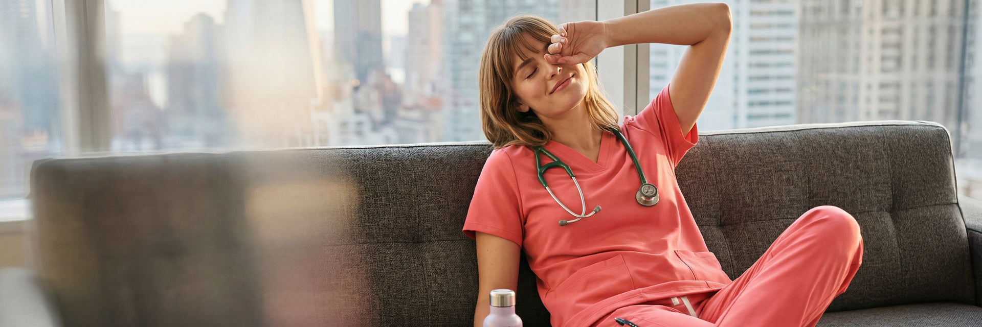A female healthcare professional in coral pink scrubs relaxing in a lounge area, leaning back comfortably