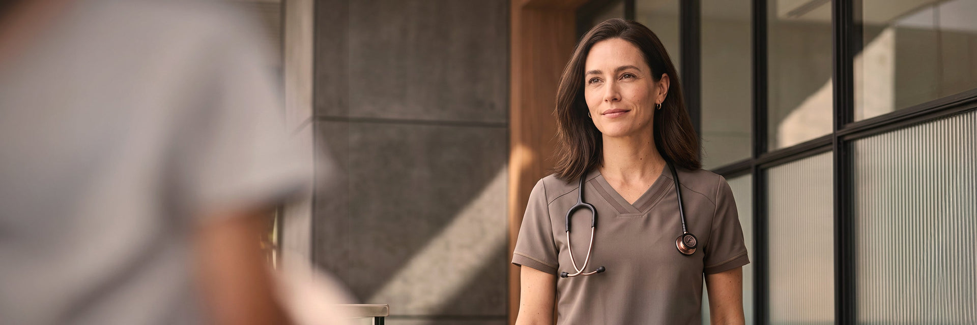 A female healthcare professional in warm-toned scrubs walking through a facility entrance, holding the door open