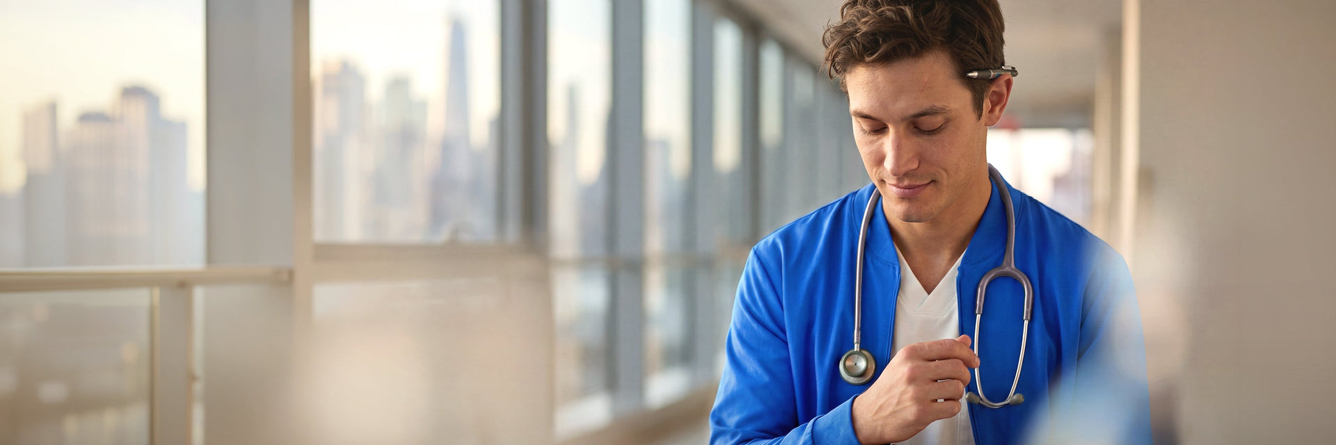 A male healthcare professional in blue scrubs walking through a modern hospital corridor, looking confident and ready for work