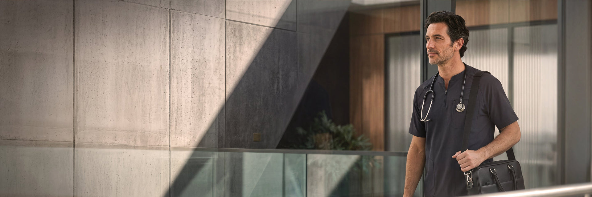 A male healthcare professional in dark charcoal scrubs standing near a glass wall in a clinical setting