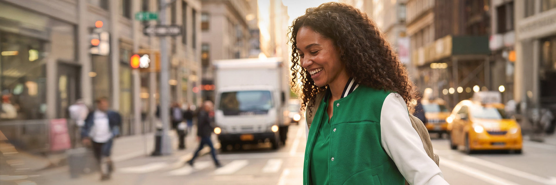 A female healthcare professional in green scrubs walking confidently on a city sidewalk with urban scenery behind her