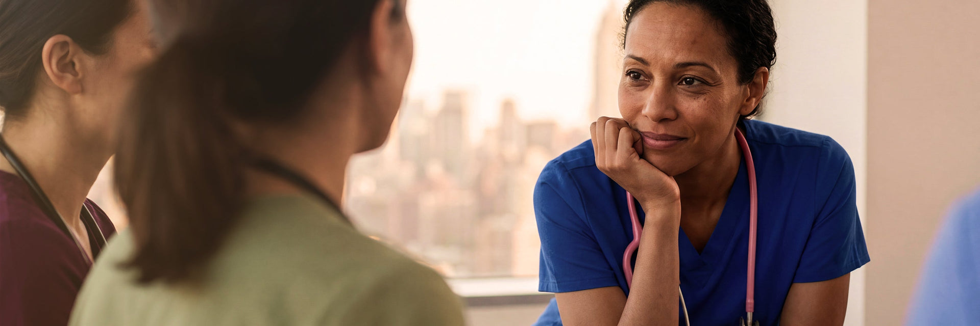 A female healthcare professional in olive green scrubs seated and speaking with a colleague in a modern lounge setting